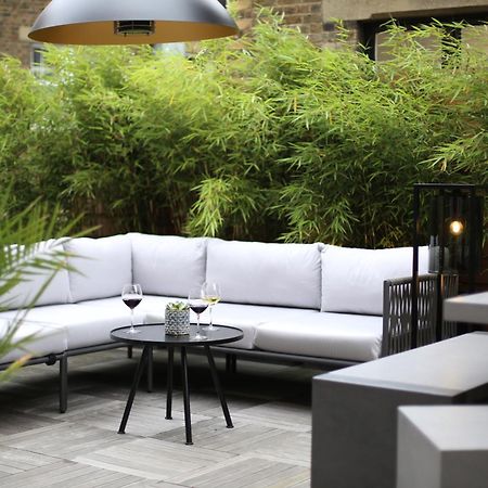 Middle Eight - Covent Garden - Preferred Hotels And Resorts 런던 외부 사진 The photo shows an outdoor seating area featuring a modern gray sectional sofa with plush cushions. In front of the sofa is a small, round black table with two glasses of wine on it, accompanied by a decorative plant or vase. Surrounding the area are